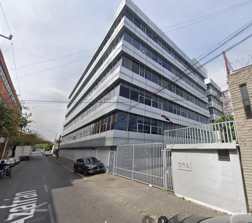 Fotografía en la que se muestra un edificio alto con ventanales. Del lado derecho del edificio hay una reja de color gris. Al terminar la reja hay un muro, también de color gris, que tiene el número 386.